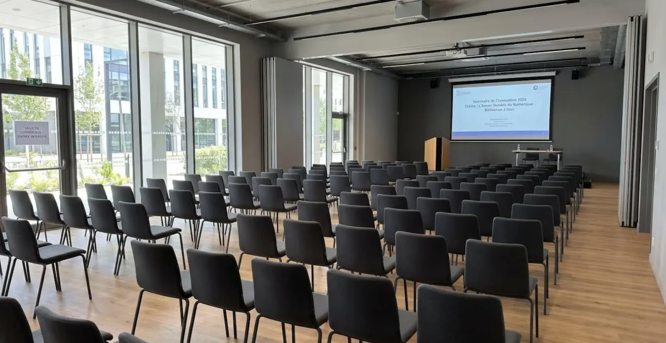 Salle de séminaire vide aménagée en configuration théâtre avec rangées de chaises alignées face à une scène et un écran de projection